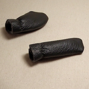 May include: Two black leather shift knob covers. The covers have a cylindrical shape with a textured surface and a stitched opening at one end. They are designed to protect the shift knob from wear.