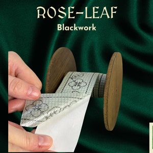 May include: A roll of white fabric with a blackwork design of flowers on a green grid. The fabric is being unrolled from a wooden spool. The text "ROSE-LEAF Blackwork" is visible above the spool.