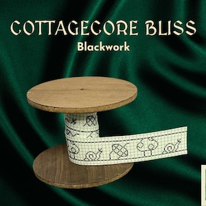 Puede incluir: Un carrete de madera con una cinta blanca con un diseño de bordado negro. La cinta tiene un patrón repetitivo de formas geométricas y árboles. El carrete está sobre un fondo de tela verde. El texto "COTTAGECORE BLISS Blackwork" está en la parte superior de la imagen.