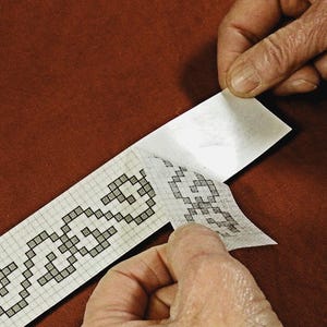 May include: A white sheet of paper with a black and white pixelated design is being peeled off of a backing sheet. The design is a repeating pattern of geometric shapes.