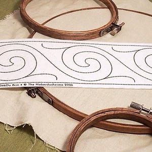 Op de afbeelding: Een witte stoffen strook met een herhalend zwart spiraalgolfpatroon, geplaatst over twee ronde houten borduurringen en een stuk beige stof. De strook bevat de gedrukte tekst: Dublin 8cm • BB046Du 8cm • © The Haberdasheress 2026.