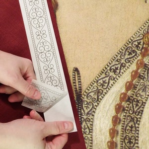 May include: A person peeling a decorative, self-adhesive border with a gray scroll design from its backing. The border is being applied to a burgundy surface, next to a section of a Renaissance-style portrait.
