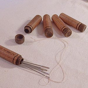 Op de afbeelding: Een set van vijf houten naaldkokers, waarvan er één open is om een ​​bundel zilveren naalden te laten zien. De kokers zijn bruin met een donkerdere bruine band rond het midden.