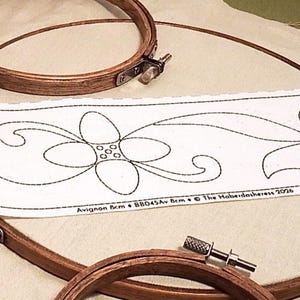 Op de afbeelding: Drie ronde houten borduurringen met metalen spanschroeven, gerangschikt op een lichtbeige stof. Een witte papieren strook bevat een zwarte lijntekening van een bloemmotief met de tekst Avignon 8cm BB045Av 8cm © The Haberdasheress 2026.