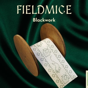 Op de afbeelding: Een houten spoel met een wit lint met een zwartwerk ontwerp van aardbeien en geometrische patronen. De tekst "FIELDMICE Blackwork" staat boven de spoel.