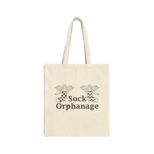 May include: A beige tote bag with black handles and a graphic of two angels wearing socks with the text "Sock Orphanage".