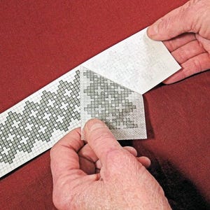 Op de afbeelding: Een close-up van een kruissteekpatroon dat van een achtergrond wordt afgepeld. Het patroon is een herhalend geometrisch ontwerp in grijstinten op een witte achtergrond. De achtergrond is een effen rode stof.