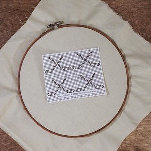 Peut inclure: Un cercle à broder en bois maintient un tissu avec un motif imprimé de quatre crosses de hockey croisées. Le motif est sur une grille, et le texte "Hockey Sticks © 2026 The Haberdasheress" est imprimé en dessous.