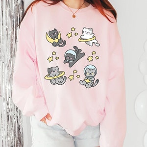 Peut inclure: Sweat-shirt rose clair avec un motif ludique de chats de dessin anim&eacute; en combinaisons spatiales et flottant dans l'espace. Les chats sont entour&eacute;s d'&eacute;toiles. Le texte "Light Pink" est en bas &agrave; gauche.