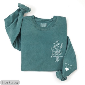 May include: A teal blue long-sleeved t-shirt with a white floral design and the text "grandma" on the front. The left sleeve has the text "james emma" and a heart.