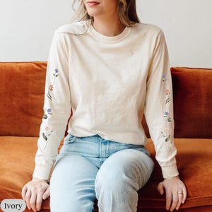 May include: An ivory long-sleeve crewneck shirt featuring delicate floral embroidery running down the length of both sleeves. The embroidery includes small stems with blue, orange, and pink flowers. The shirt is paired with light blue denim jeans.