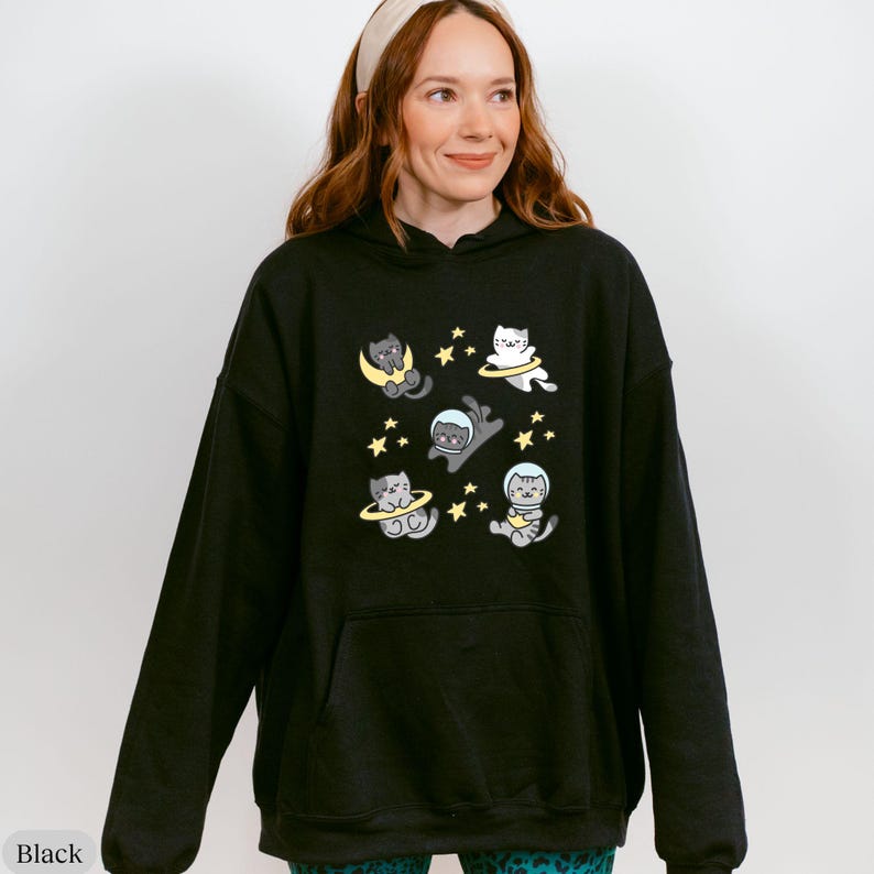 Peut inclure: Sweat &agrave; capuche noir avec un motif fantaisiste de chats de dessin anim&eacute; dans l'espace, des lunes et des &eacute;toiles. Le motif est en gris, jaune et blanc. Le sweat &agrave; capuche a une capuche &agrave; cordon et une poche kangourou.