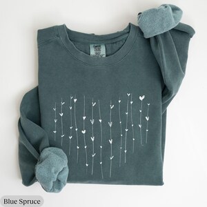 May include: A teal blue sweatshirt with a white heart design. The design features a series of vertical lines with small hearts along each line. The sweatshirt has a crew neck and long sleeves.