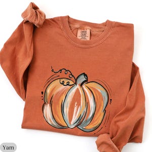 May include: A long-sleeve crewneck sweatshirt in a muted burnt orange color. The front features a stylized graphic of a pumpkin painted with brushstrokes of orange, cream, and sage green. The inner neck label reads Comfort Colors.
