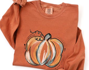 May include: A long-sleeve crewneck sweatshirt in a muted burnt orange color. The front features a stylized graphic of a pumpkin painted with brushstrokes of orange, cream, and sage green. The inner neck label reads Comfort Colors.