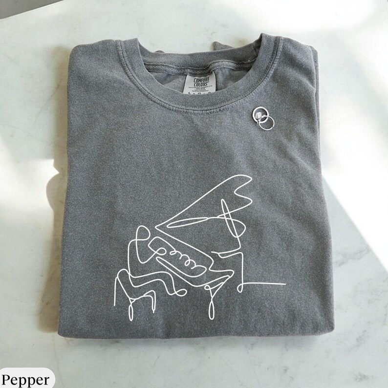 Pode incluir: Uma camiseta cinza-chumbo com um desenho de linha branca de uma pessoa tocando piano. A camiseta tem uma etiqueta que diz "Comfy Colors".