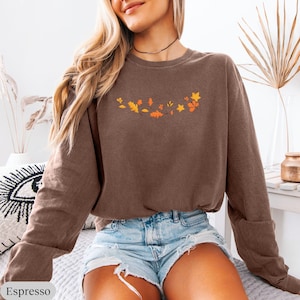 May include: A long-sleeve crewneck sweatshirt in a solid espresso brown color. The chest area features a small, centered graphic print consisting of various autumn leaves in shades of yellow, orange, and red, arranged in a horizontal arc.