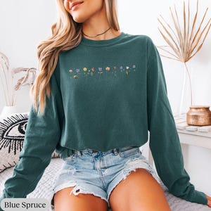 Op de afbeelding: Een tealblauwe longsleeve met een rij kleurrijke geborduurde bloemen over de borst. De trui wordt gedragen door iemand die een spijkerbroek draagt.