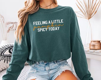 Może przedstawiać: Ciemnozielona bluza z długim rękawem i okrągłym dekoltem, z białym i pomarańczowym napisem "FEELING A LITTLE pumpkin SPICY TODAY" nadrukowanym na klatce piersiowej. Odzież ma luźny krój z prążkowanymi mankietami i wykończeniem dekoltu.