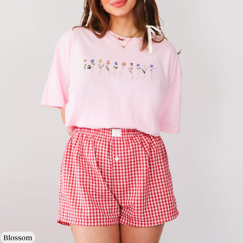 Op de afbeelding: Een roze T-shirt met een rij geborduurde bloemen over de borst. Het T-shirt wordt gecombineerd met rood-wit geblokte shorts.