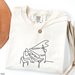 Pode incluir: Uma camiseta branca com um desenho de linha preta de uma pessoa tocando piano. A camiseta tem uma etiqueta que diz "Comfort Colors".