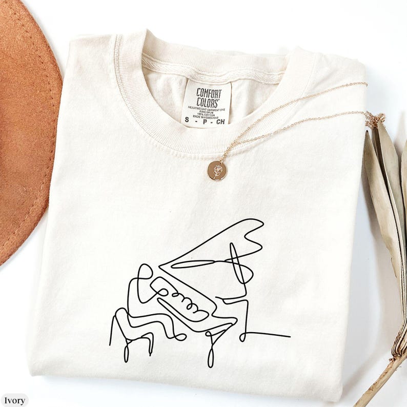 Pode incluir: Uma camiseta branca com um desenho de linha preta de uma pessoa tocando piano. A camiseta tem uma etiqueta que diz "Comfort Colors".