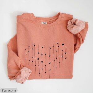 May include: Terracotta colored sweatshirt with a black heart design made of lines and small hearts. The sweatshirt has a crew neck and long sleeves.