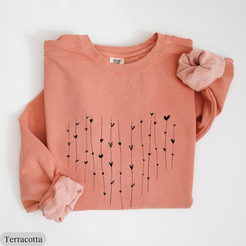 May include: Terracotta colored sweatshirt with a black heart design made of lines and small hearts. The sweatshirt has a crew neck and long sleeves.