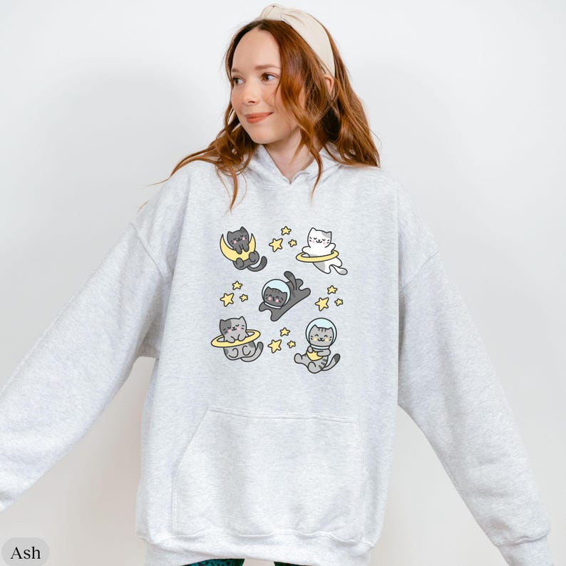 Peut inclure: Sweat &agrave; capuche gris clair avec un motif amusant de chats de dessin anim&eacute; en combinaisons spatiales, flottant parmi les &eacute;toiles et les corps c&eacute;lestes. Le motif comprend des chats sur des lunes et dans des anneaux spatiaux. Le mot "Ash" est imprim&eacute; en bas &agrave; gauche.