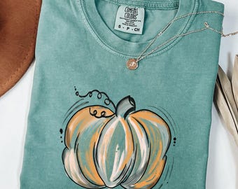 May include: Light green Comfort Colors t-shirt with a hand-drawn pumpkin graphic. The pumpkin is orange, white, and black. A gold necklace with a small pendant is on the shirt. The shirt is labeled "S P CH".