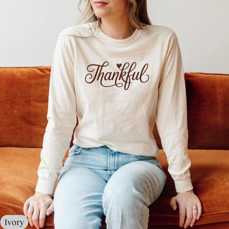 May include: Ivory long-sleeve shirt with the word "Thankful" in a brown script font. The word "Thankful" has a small heart above the "f". The shirt is paired with blue jeans.