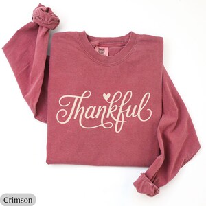 May include: Crimson long-sleeve shirt with the word "Thankful" in a cursive font and a small heart. The shirt is made of a soft material and has a crew neck. The sleeves are cuffed at the wrists. The shirt is a solid crimson color.