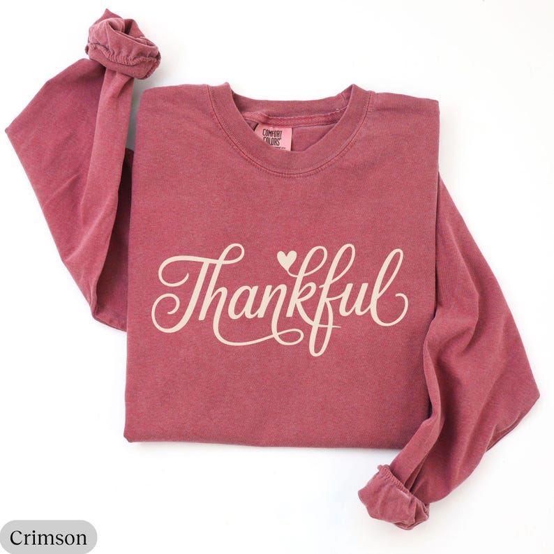 May include: Crimson long-sleeve shirt with the word "Thankful" in a cursive font and a small heart. The shirt is made of a soft material and has a crew neck. The sleeves are cuffed at the wrists. The shirt is a solid crimson color.