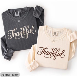 May include: Two long-sleeved shirts, one charcoal gray and one ivory, are displayed. The word "Thankful" is scripted across the front of each shirt. A small heart is above the "ful". Each shirt has a crew neck and a small pendant necklace.