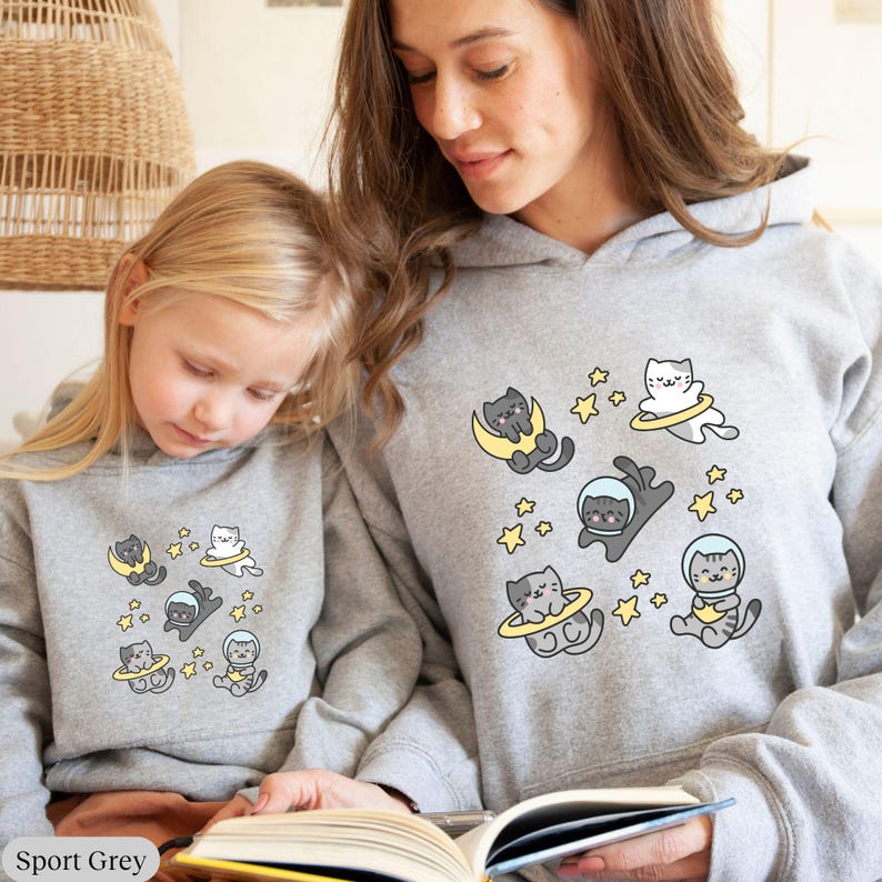 Peut inclure: Deux sweats &agrave; capuche gris avec un motif sur le th&egrave;me des chats. Le motif pr&eacute;sente des chats de dessins anim&eacute;s en combinaisons spatiales, sur des lunes et avec des halos jaunes, entour&eacute;s d'&eacute;toiles jaunes. Les sweats &agrave; capuche sont &eacute;tiquet&eacute;s "Sport Grey" en bas.