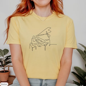 Pode incluir: Uma camiseta amarela com um desenho de linha preta de uma pessoa tocando piano.