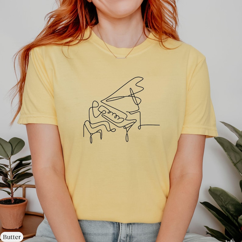 Pode incluir: Uma camiseta amarela com um desenho de linha preta de uma pessoa tocando piano.