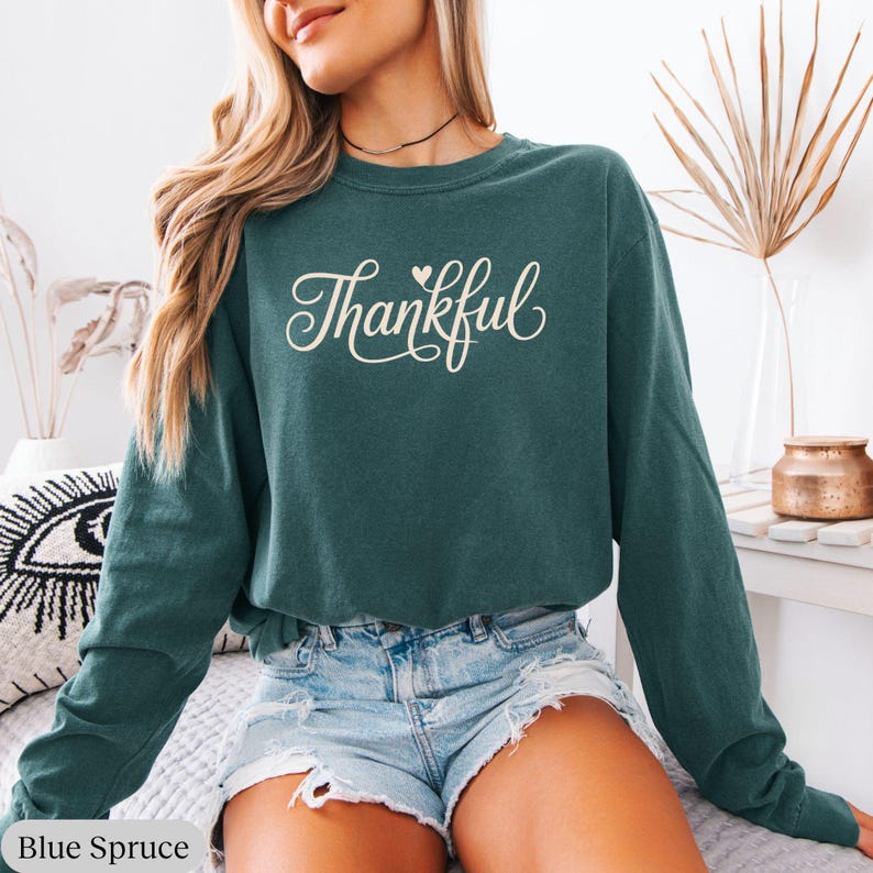 May include: A long-sleeved, crew neck sweatshirt in blue spruce color. The word "Thankful" is written in a cursive font across the front. The sweatshirt is paired with denim shorts.