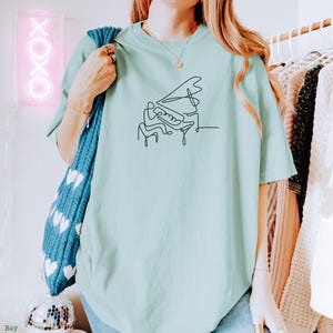 Pode incluir: Uma camiseta azul clara com um desenho de linha preta de uma pessoa tocando piano.