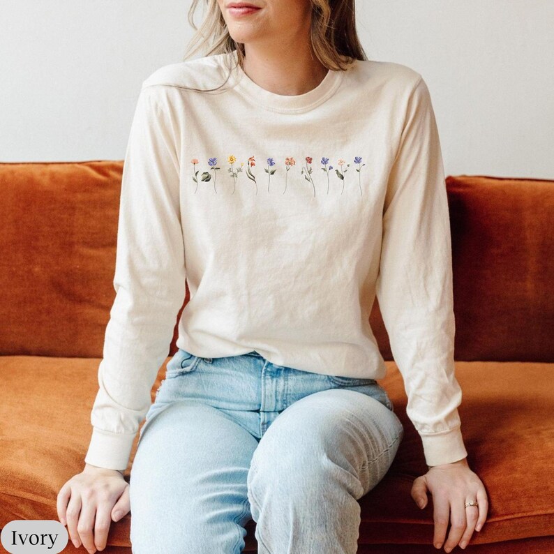 Op de afbeelding: Een langarm, ivoorwit T-shirt met een rij van kleurrijke geborduurde bloemen over de borst.