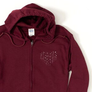 May include: A maroon zip-up hoodie with a white heart design made of small hearts and stems. The hoodie has a drawstring hood.