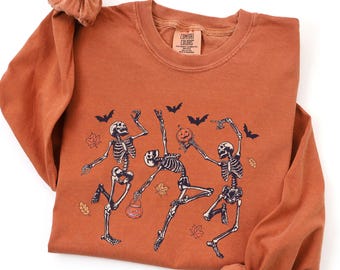 May include: A long-sleeve crewneck sweatshirt in a burnt orange color featuring a graphic print of three dancing skeletons, bats, and autumn leaves. One skeleton holds a jack-o'-lantern. The inner neck label reads Comfort Colors.