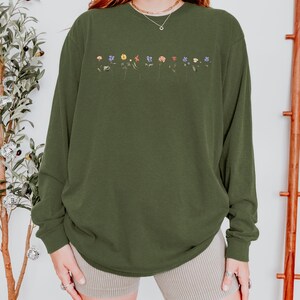Op de afbeelding: Een groen shirt met lange mouwen met een rij geborduurde bloemen in verschillende kleuren over de borst. Het shirt is gemaakt van een zachte, comfortabele stof en heeft een ontspannen pasvorm.