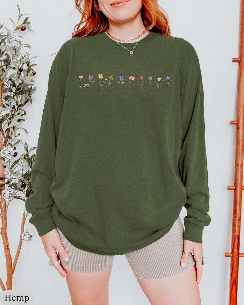 Op de afbeelding: Een groen shirt met lange mouwen met een rij geborduurde bloemen in verschillende kleuren over de borst. Het shirt is gemaakt van een zachte, comfortabele stof en heeft een ontspannen pasvorm.