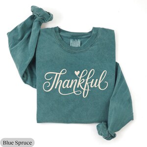 May include: A teal-colored sweatshirt with the word "Thankful" in a cursive font, with a small heart above the "f". The sleeves are bunched up. The sweatshirt is made of a soft material.