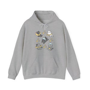Peut inclure: Un sweat &agrave; capuche gris clair orn&eacute; d'un motif ludique de chats de dessin anim&eacute; en combinaisons spatiales et flottant dans l'espace. Les chats sont entour&eacute;s d'&eacute;toiles et de plan&egrave;tes. Le sweat &agrave; capuche a une poche avant et une capuche &agrave; cordon.
