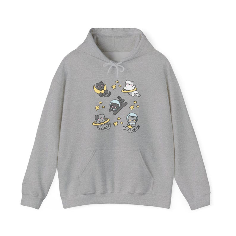 Peut inclure: Un sweat &agrave; capuche gris clair orn&eacute; d'un motif ludique de chats de dessin anim&eacute; en combinaisons spatiales et flottant dans l'espace. Les chats sont entour&eacute;s d'&eacute;toiles et de plan&egrave;tes. Le sweat &agrave; capuche a une poche avant et une capuche &agrave; cordon.