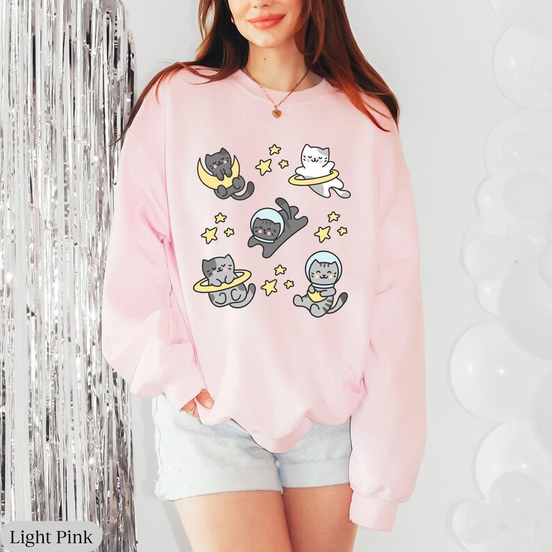 Peut inclure: Sweat-shirt rose clair avec un motif ludique de chats de dessin anim&eacute; en combinaisons spatiales et flottant dans l'espace. Les chats sont entour&eacute;s d'&eacute;toiles. Le texte "Light Pink" est en bas &agrave; gauche.