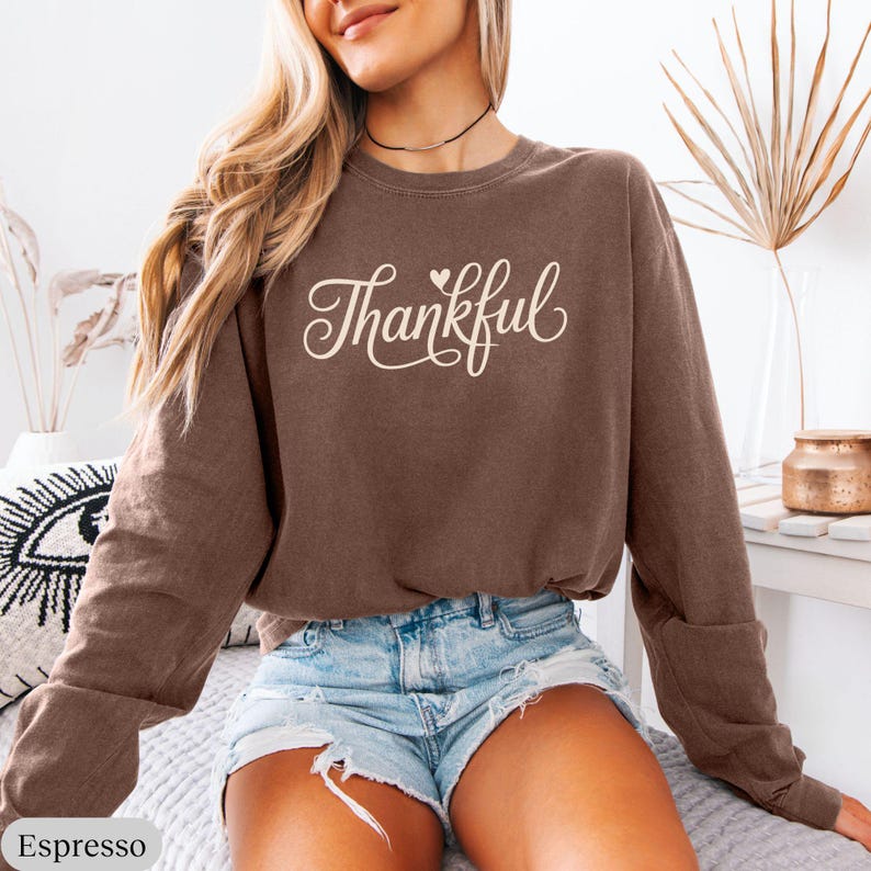 May include: Espresso-colored sweatshirt with the word "Thankful" in a cursive font. The long-sleeved top has a crew neck and is paired with denim shorts. The sweatshirt is a solid color and has a relaxed fit.
