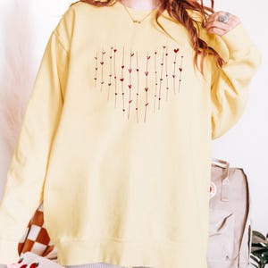 May include: A yellow sweatshirt with a red heart design made of small hearts and lines. The sweatshirt is oversized and has a crew neck.